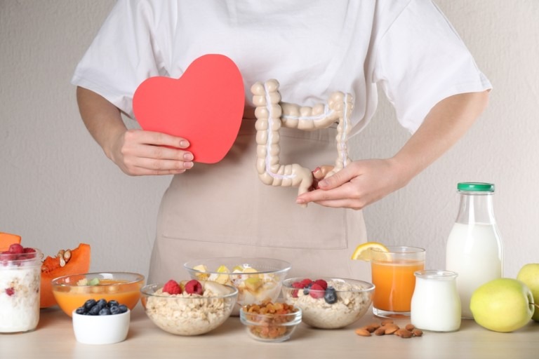 Foto de uma mulher, que não aparece seu rosto, ela segura um coração vermelho de papel e um objeto que imita o intestino. Há uma mesa cheia de recipientes com sucos e comidas.